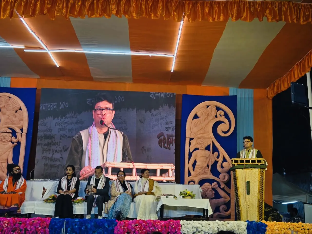 Dr. Jayanta Ghosh addressing the Boi Mela 2026 audience from the podium with a large screen projection behind him