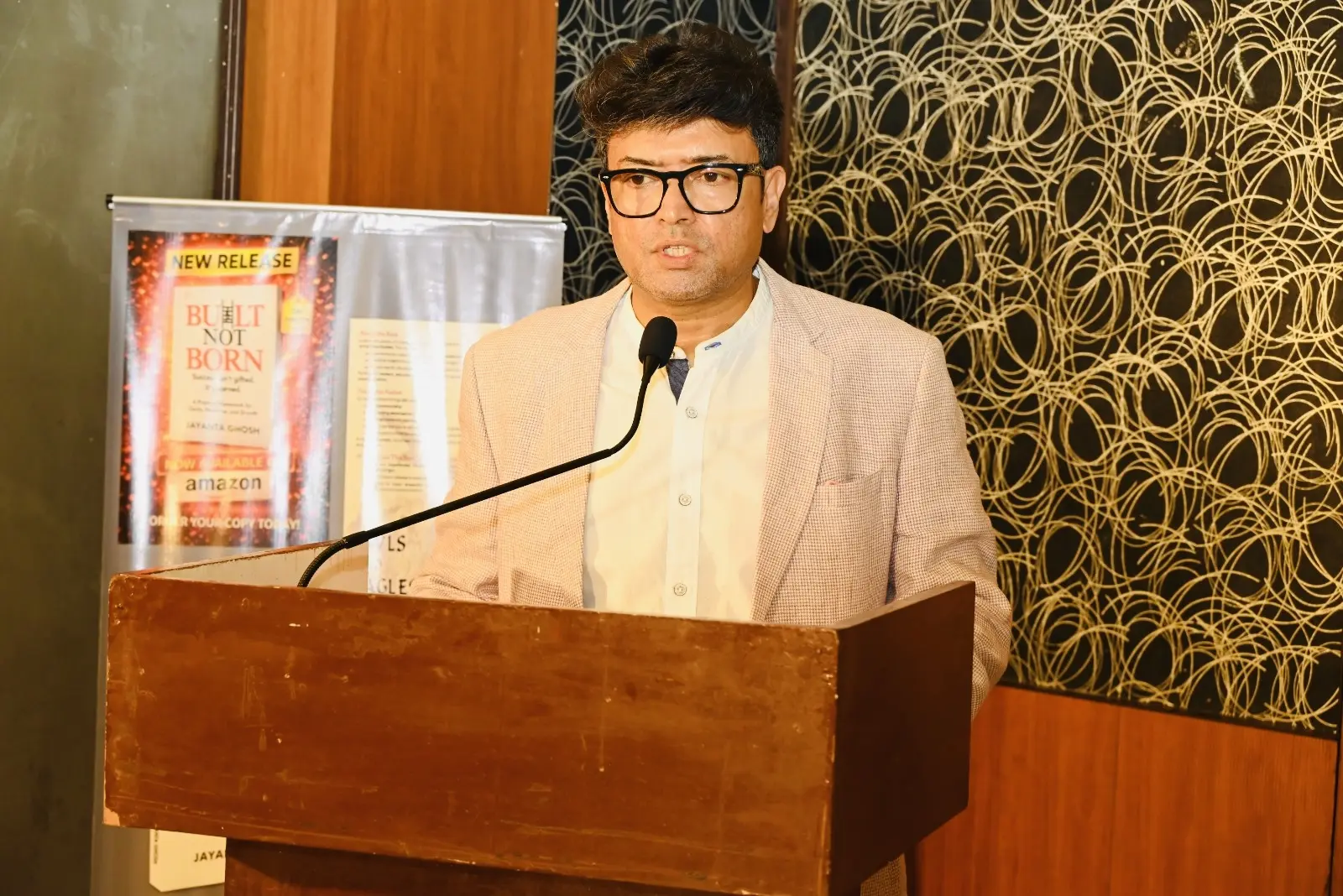 Dr. Jayanta Ghosh speaking at a podium beside the Built Not Born new release banner at the book launch