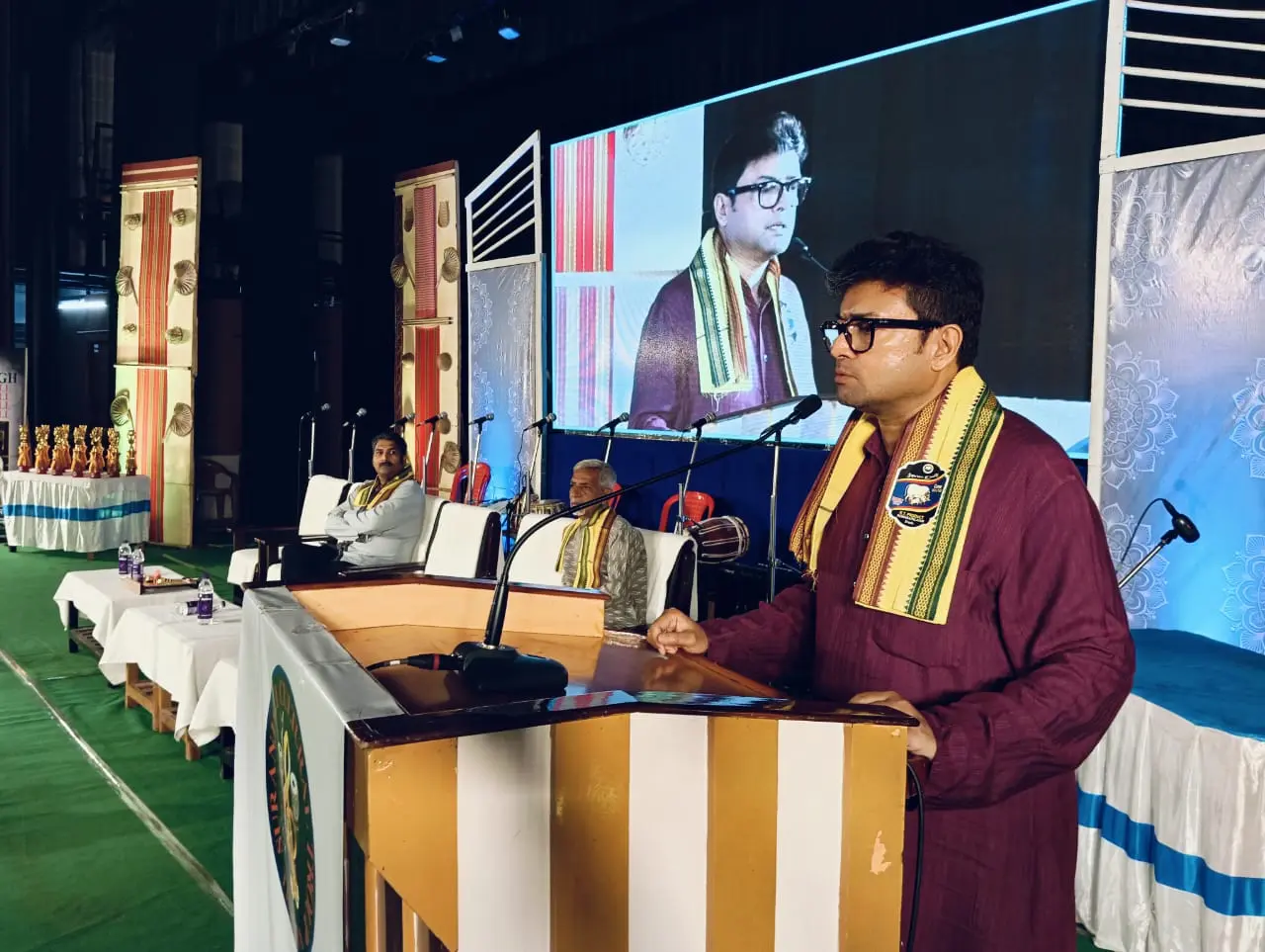 Dr. Jayanta Ghosh delivering the Folk Utsav keynote from the podium with a large LED screen behind the stage
