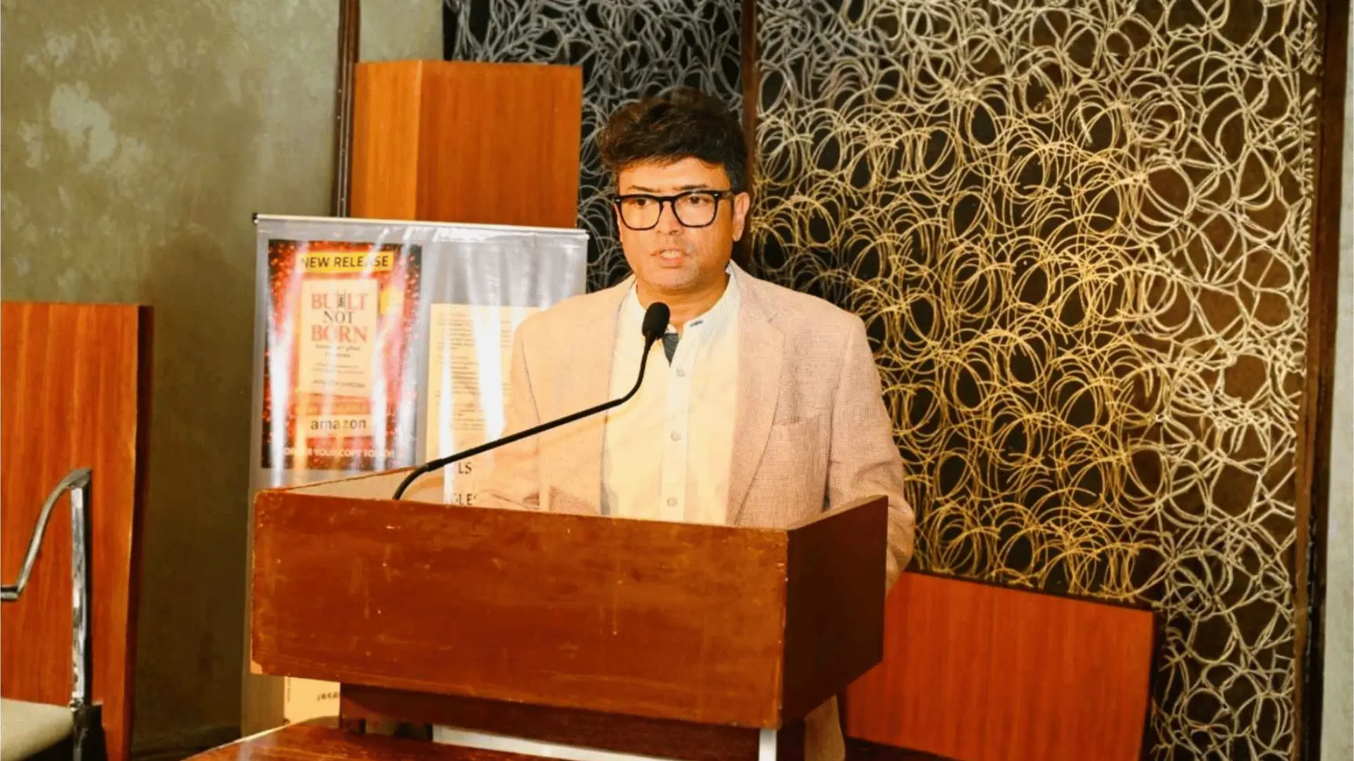Dr. Jayanta Ghosh speaking at a podium beside the Built Not Born book launch banner